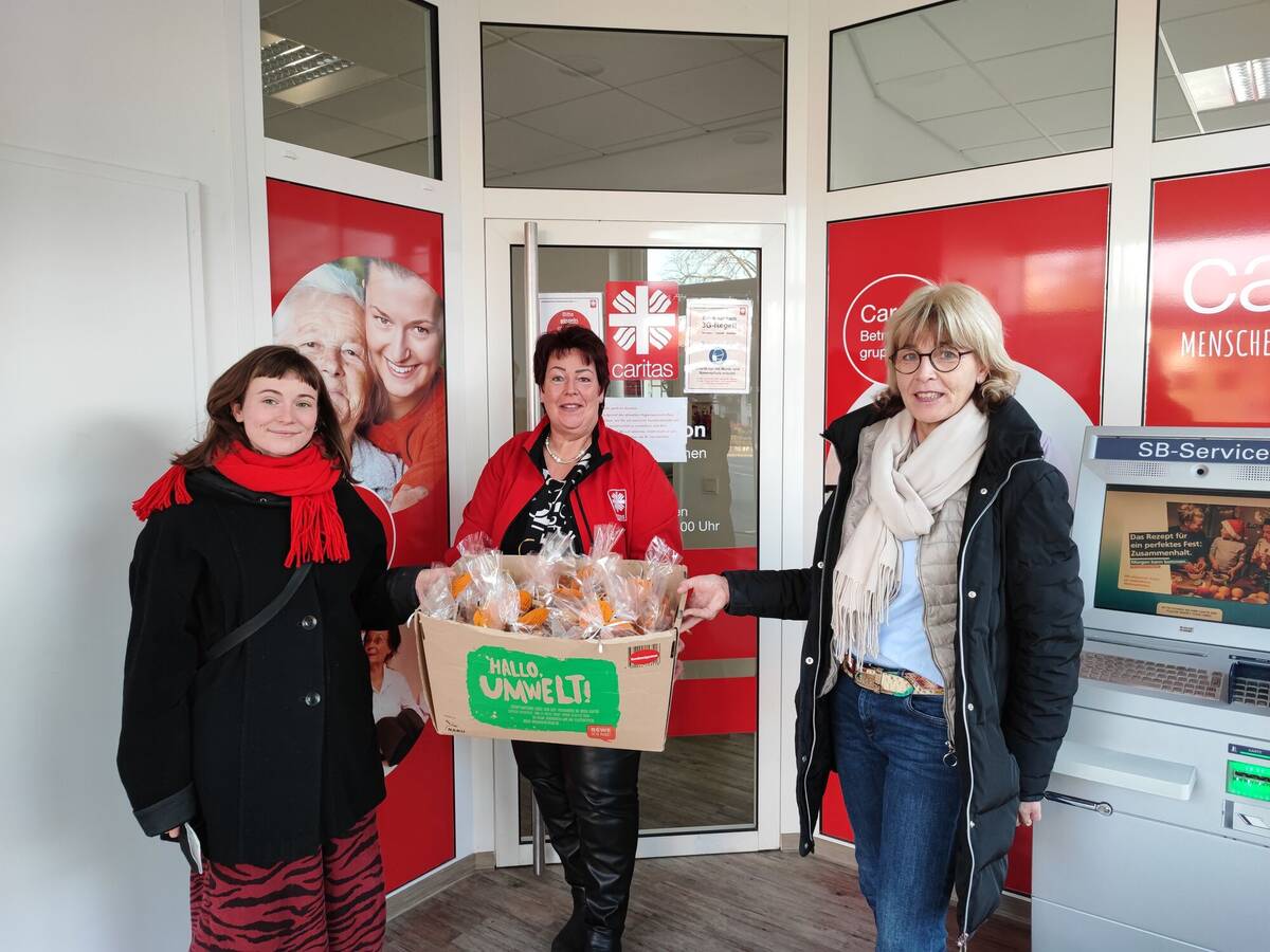 Tabea Below (l.) und Daniela Ramb (r.) aus der khg übergeben die gepackten Plätzchentüten vor der Sozialstation an Christiane Koch von der Caritas.